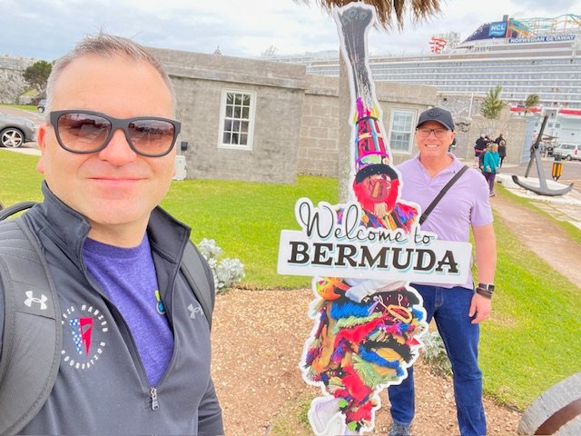 Dois homens tirando uma selfie de um "Bem vindo às Bermudas" placa com um prédio de pedra cinza e um navio de cruzeiro ao fundo.