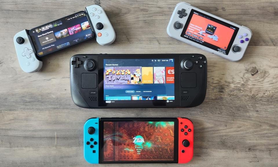 Uma coleção de dispositivos portáteis de jogos descansa em uma mesa de madeira. Os dispositivos incluem o Nintendo Switch - Modelo OLED, Valve Steam Deck e o Retroid Pocket 3, além de um iPhone 12 mini conectado a um controlador de jogos móvel Backbone One.