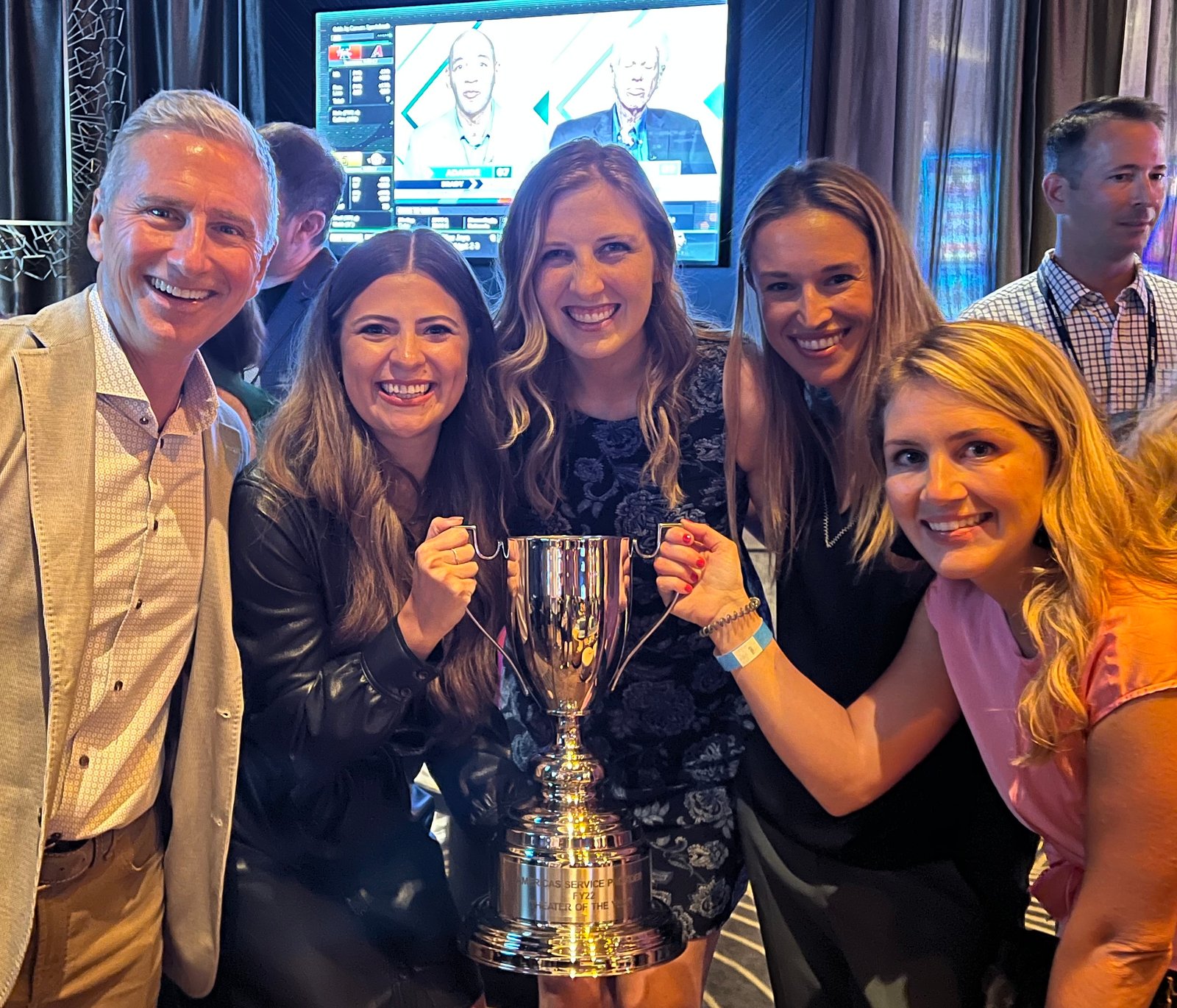 Um homem e quatro mulheres posam juntos segurando um grande troféu de prata.